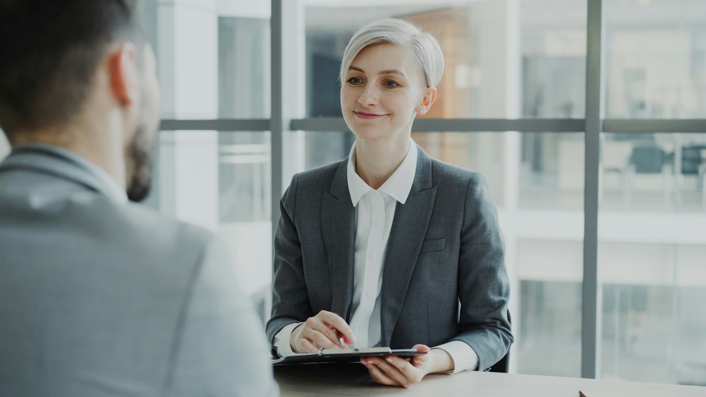Donna in tailleur grigio intervista un uomo seduto alla scrivania in un ufficio moderno.